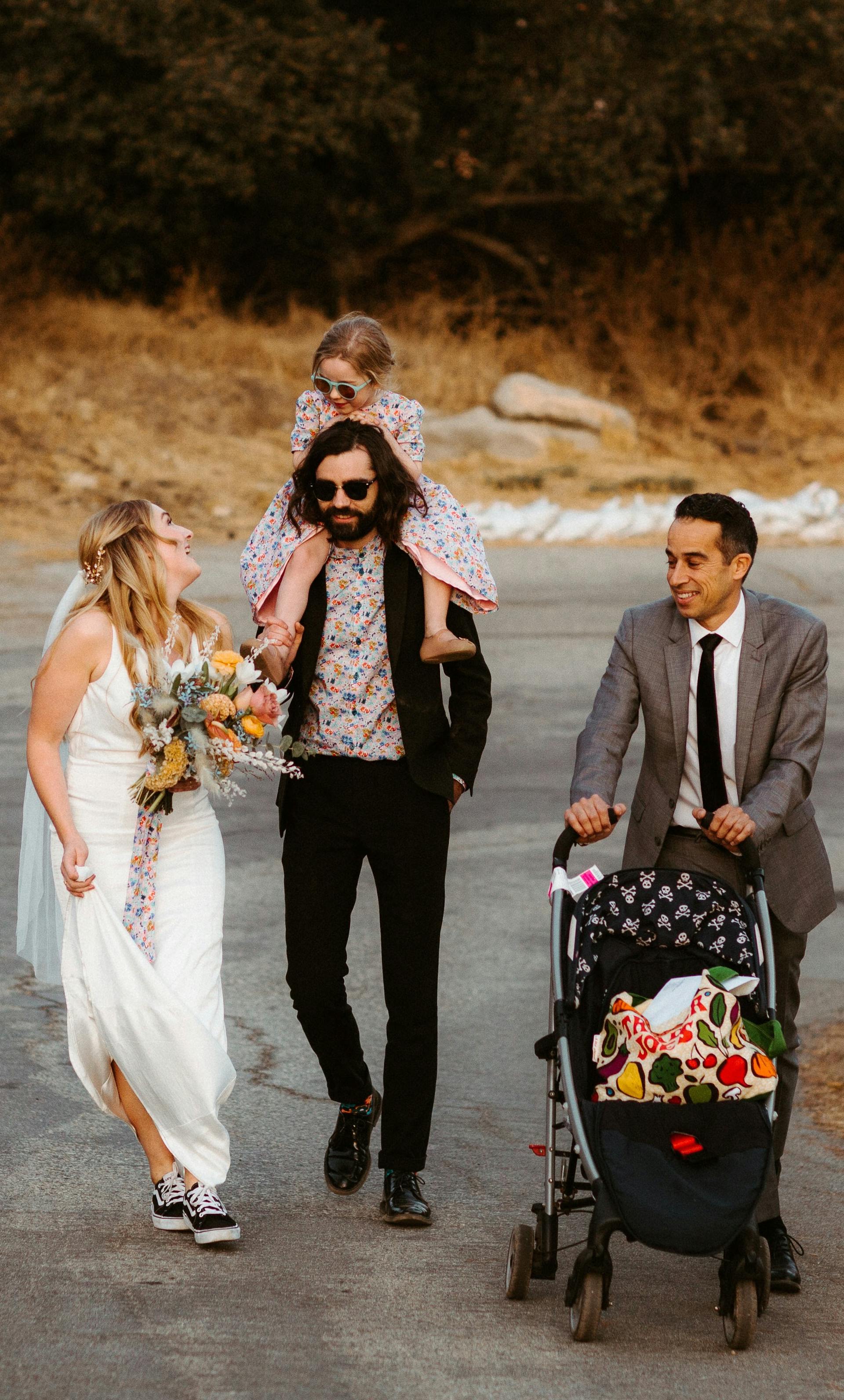 a photo of jacob pushing a stroller up a hill as the couple walk next to him.  There is a little girl sitting on the shoulder of the groom
